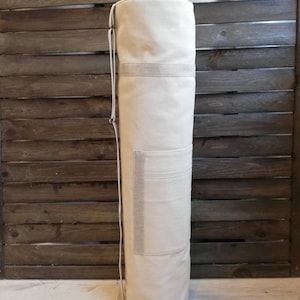 K&ouml;nnte beinhalten: Eine wei&szlig;e Yogamatte aus Canvas mit Kordelzug und einer kleinen Tasche an der Seite. Die Tasche steht aufrecht vor einem Holz Hintergrund.