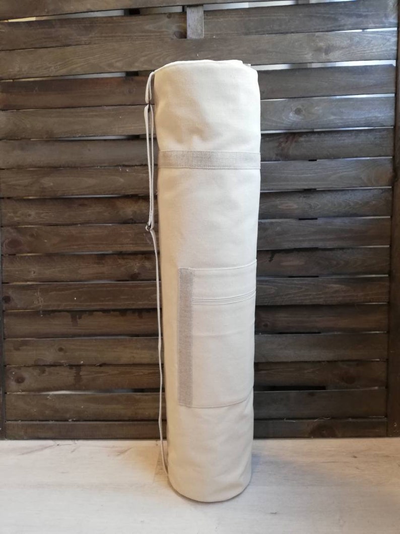 K&ouml;nnte beinhalten: Eine wei&szlig;e Yogamatte aus Canvas mit Kordelzug und einer kleinen Tasche an der Seite. Die Tasche steht aufrecht vor einem Holz Hintergrund.