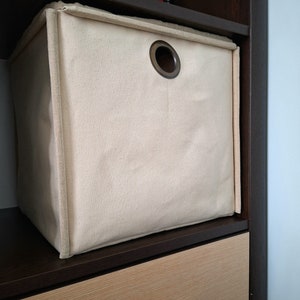 Peut inclure: Un bac de rangement en toile beige avec une poignée en métal brun. Le bac est posé sur une étagère brun foncé avec un tiroir brun clair en dessous.