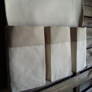 Könnte beinhalten: Drei weiße Canvas-Aufbewahrungstaschen mit braunen Stoffoberteilen, die an einer Holzwand hängen.