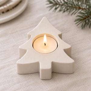 May include: A cream-colored, tree-shaped candle holder made of a smooth, matte material. The holder features a central circular cutout containing a small, lit tea light candle with a visible flame, creating a warm glow within the structure.