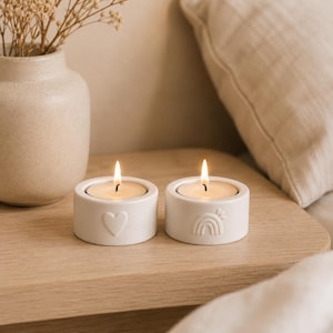 May include: Two white ceramic tea light candle holders with a cylindrical shape. One holder features an embossed heart design, while the other displays an embossed rainbow motif. Each holder contains a lit tea light candle with a visible flame.