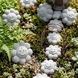 Peut inclure: Un jardin miniature dans un pot en céramique rond comprenant plusieurs pierres de jardin en béton gris clair en forme de fleurs avec des visages souriants, une petite maison en brindilles, une arche en métal bronze, de la mousse et des plantes grasses.