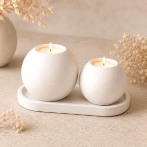 Może przedstawiać: Dwa kuliste, białe świeczniki o matowym wykończeniu, z których każdy zawiera zapaloną świeczkę typu tealight. Świeczniki są ustawione na owalnej, białej tacy, tworząc spójny zestaw. Powierzchnia świeczników i tacy jest gładka i jednolita.