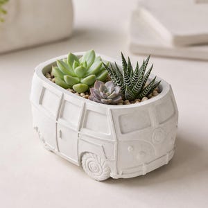 Op de afbeelding: Een witte betonnen plantenbak in de vorm van een vintage camperbusje, met drie kleine vetplantjes in lichte steentjes. De plantenbak heeft gedetailleerde vormen van ramen, wielen en een voorbumper, wat zorgt voor een textuurrijk oppervlak.