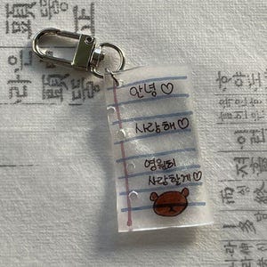 Könnte beinhalten: Ein klarer Acryl-Schlüsselanhänger in Notizblockform mit silberfarbener Hardware. Der Notizblock hat Linien und handschriftlichen koreanischen Text, einschließlich einer kleinen braunen Bärenillustration. Der Schlüsselanhänger ist an einem silbernen Verschluss befestigt.