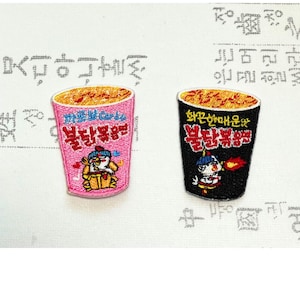 Könnte beinhalten: Zwei gestickte Aufnäher in Form von Instant-Nudelbechern. Einer ist rosa, der andere schwarz. Beide haben orangefarbene Nudeln und koreanischen Text. Der rosa Becher zeigt eine Zeichentrickfigur. Der schwarze Becher hat den Text "화끈한매운맛" und eine Zeichentrickfigur.
