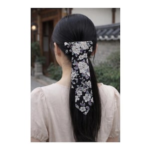May include: A black hair bow featuring a floral design with white and purple flowers. The bow is tied and hangs down the back of the head. The hair accessory is worn by a person with long, dark hair.