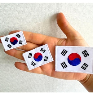 Op de afbeelding: Drie witte rechthoekige patches met het ontwerp van de Zuid-Koreaanse vlag. Elke patch heeft een witte rand, een centraal rood en blauw Taegeuk-symbool en vier zwarte trigrammen. De patches worden in een hand gehouden.