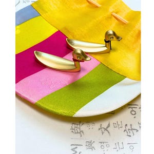 Peut inclure: Deux objets dorés en forme de canard avec des accents rouges et bleus, posés sur un tissu coloré avec des sections roses, jaunes et vertes. Les canards ont un design épuré et minimaliste.