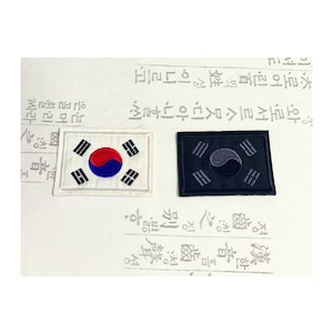 Op de afbeelding: Twee geborduurde patches met de Zuid-Koreaanse vlag, de Taegeukgi. Eén patch is wit met een rode en blauwe Taegeuk en zwarte trigrammen, de andere is zwart met een grijze Taegeuk en trigrammen.