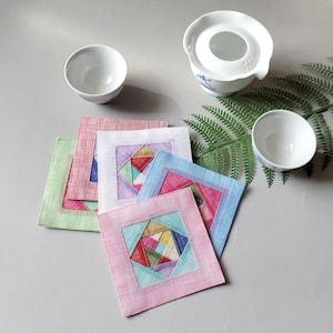 Pode incluir: Uma coleção de bases quadradas de tecido colorido com desenhos geométricos, dispostas com chávenas de chá e uma bule de cerâmica brancos. As bases apresentam cores pastel, rosa, azul e verde, numa superfície cinzenta.