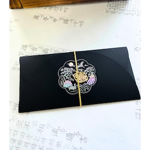 Op de afbeelding: Een zwarte envelop met een decoratief embleem met een kraanvogel, bergen en een goudkleurig symbool. De envelop is vastgebonden met een gouden touwtje. De achtergrond is een wit oppervlak met Koreaanse tekst.