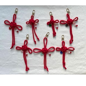 Puede incluir: Varios llaveros rojos con nudos chinos y cierres dorados. Cada llavero presenta un diseño de nudo decorativo con cordones colgantes y pequeñas cuentas de colores. Los llaveros están dispuestos sobre una superficie blanca.