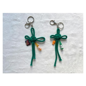 Puede incluir: Dos llaveros de cuerda verde con cierres plateados. Cada llavero presenta un nudo decorativo y pequeños dijes de botella coloridos. Un llavero tiene una pequeña etiqueta. El fondo es una superficie blanca texturizada.