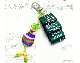 Może przedstawiać: Kolorowy koreański tradycyjny brelok z amuletem szczęścia. Urok ma pasiasty, pękaty korpus w kolorze czerwonym, niebieskim, zielonym i różowym, z zielonym węzłem i frędzlami. Zielona wstążka z białym tekstem jest przymocowana do srebrnego zapięcia.