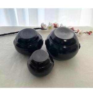 Puede incluir: Tres ollas de cerámica negra de diferentes tamaños están dispuestas sobre una superficie blanca. Las ollas tienen forma redondeada y un acabado brillante. Ramas florales en el fondo.