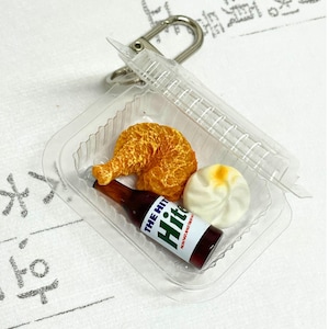 Puede incluir: Llavero de comida en miniatura con una pata de pollo frita, un bollo blanco con la parte superior amarilla y una botella marrón con el texto "THE HIT". Los alimentos están en un recipiente de plástico transparente, sujeto a un llavero plateado.