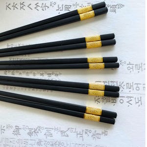 May include: Black chopsticks with gold bands. The chopsticks are arranged in pairs, with a gold band near the top of each pair. The background is a white surface with Korean text.