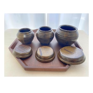 May include: A set of three brown ceramic pots with lids, displayed on a wooden tray. The pots vary in size, with a dark interior and a textured exterior. The lids are round and match the pots' color and texture.