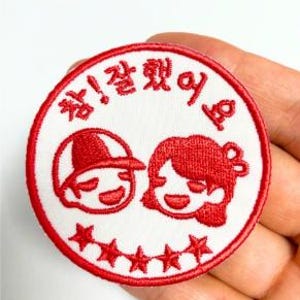 Puede incluir: Un parche circular blanco con detalles bordados en rojo. El parche presenta dos figuras de dibujos animados sonrientes y el texto coreano "참! 잘했어요" en rojo. Debajo de las figuras hay seis estrellas rojas y el parche tiene un borde rojo.