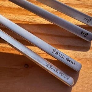 Może przedstawiać: Zbliżenie czterech srebrnych pałeczek z tekstem. Pałeczki są ułożone po przekątnej na drewnianej powierzchni. Tekst na pałeczkach wydaje się być w języku koreańskim i angielskim.