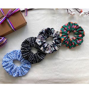 May include: Four fabric scrunchies in a row. The scrunchies have different patterns: blue floral, black with white lines, black with white flowers, and green with red and orange shapes. A brown gift box with a colorful ribbon is in the background.