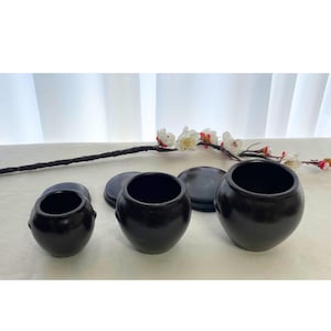 May include: Three black ceramic jars of varying sizes, each with a matching lid. The jars have a rounded shape with a wide opening. A branch with white flowers and red accents is in the background.