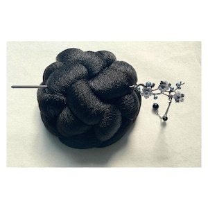 May include: A black hair bun with a decorative hairpin. The hairpin features a floral design with small blue and silver accents and dangling beads. The bun is made of braided hair and is a dark, solid color.
