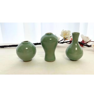 Puede incluir: Se muestran tres pequeños jarrones de cerámica verde salvia de diferentes formas. Los jarrones están dispuestos sobre una superficie de color claro, con una rama de flores blancas en el fondo. Los jarrones tienen un acabado brillante.
