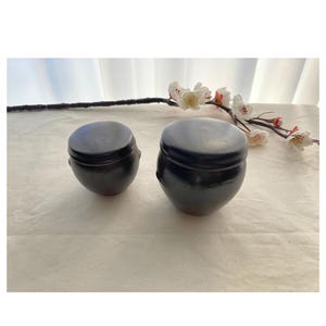 May include: Two small, dark brown ceramic jars with lids. The jars are round with a slightly bulbous shape. A sprig of white and red flowers is in the background. The jars are sitting on a white surface.