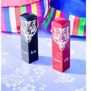 Puede incluir: Dos sellos rectangulares, uno negro y otro rojo, cada uno con un diseño de cabeza de tigre blanco. El sello negro tiene el nombre "Louise" y un carácter. El sello rojo tiene "Steven" y un carácter. Bolsas coloridas en el fondo.