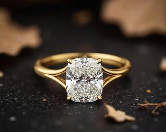 May include: A gold engagement ring featuring a large, cushion-cut diamond. The ring has a split shank design. The setting is against a dark background with blurred leaves.
