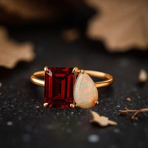 Peut inclure: Une bague en or ornée d'une grande pierre rouge foncé rectangulaire et d'une pierre blanche opalescente en forme de poire. La bague est posée sur un fond sombre, soulignant le contraste des couleurs et des textures.
