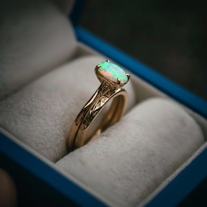 May include: A gold ring set with an oval opal gemstone, displayed in a blue ring box. The ring has an intricate floral design on the band. The opal has a play of color, with flashes of green, blue, and red.
