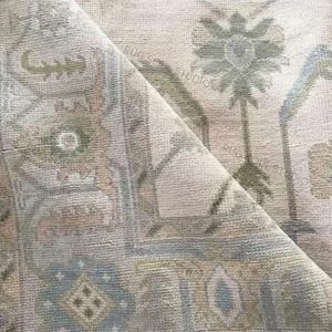 May include: A rolled-up area rug with a faded, vintage aesthetic. The rug features a complex pattern in muted tones of beige, blue, and green. The design includes floral and geometric elements, creating a traditional, yet soft look. The rug's texture is visible, suggesting a woven construction.