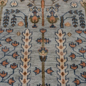 Può includere: Primo piano di un tappeto azzurro con un intricato motivo floreale. Il design presenta motivi floreali e di alberi stilizzati ripetuti in tonalità arancioni, marroni e blu. Il tappeto ha un aspetto strutturato.