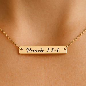 Könnte beinhalten: Eine goldfarbene Halskette mit einer feinen Kette und einem rechteckigen Anhänger. Der Anhänger ist mit den Worten "Proverbs 3:5-6" in kursiver Schrift versehen. Die Halskette wird vor einem hautfarbenen Hintergrund gezeigt.