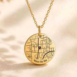 May include: A gold-toned circular pendant necklace featuring an engraved street map design with a black location pin icon. The text "Where We Met" is inscribed at the bottom of the disc, which hangs from a delicate matching gold-toned chain.