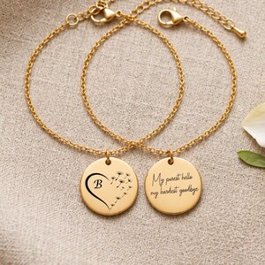 May include: Two gold-toned chain bracelets featuring circular pendants. One pendant displays a black letter B inside a heart shape formed by dandelion seeds. The other pendant is engraved with the black text, My purest hello, my hardest goodbye.