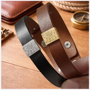 May include: Two leather bracelets, one black and one brown, each featuring a square metal slider. The black bracelet has a silver-toned slider with a black star map engraving, while the brown bracelet has a gold-toned slider with a black star map.