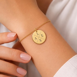 Puede incluir: Pulsera dorada con un colgante redondo. El colgante está dividido en cuatro secciones, cada una con una letra: L, E, A y X. La pulsera se lleva en la muñeca.