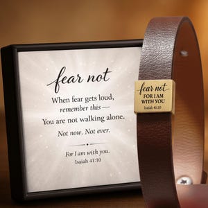 May include: A framed print and a leather bracelet. The print features the words "fear not" in elegant script, with the text "When fear gets loud, remember this - You are not walking alone. Not now. Not ever. For I am with you. Isaiah 41:10". The bracelet has a gold-coloured metal piece with the words "fear not" and "FOR I AM WITH YOU Isaiah 41:10".