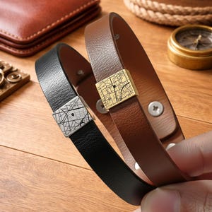 Puede incluir: Dos pulseras de cuero, una negra y otra marrón, cada una con un pasador metálico cuadrado grabado con un diseño de mapa de calles detallado. La pulsera negra tiene un pasador plateado, mientras que la marrón tiene un pasador dorado.
