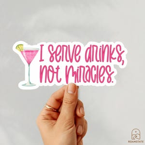 May include: A sticker with the text "I serve drinks, not miracles" in pink script. A pink cocktail with a lime wedge is on the left. The sticker is held by a hand.