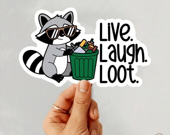May include: A sticker featuring a cartoon raccoon wearing sunglasses, holding a green trash can filled with items. The text "Live. Laugh. Loot." is written in black. The sticker is held by a hand.