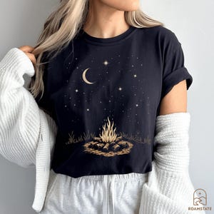 May include: Black t-shirt featuring a campfire graphic under a starry night sky with a crescent moon. The design includes a campfire with flames, logs, and surrounding grass. The shirt has short sleeves and a relaxed fit.