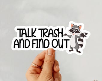 May include: A white sticker with the text "TALK TRASH AND FIND OUT" in black, featuring a cartoon raccoon. The raccoon is gray, white, and black, with its arms outstretched. The sticker is held by a hand.