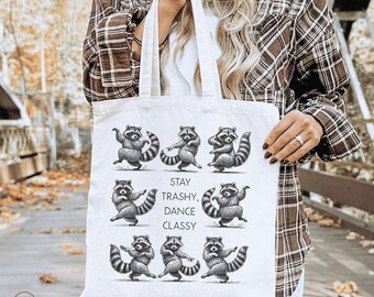 Könnte beinhalten: Eine weiße Tragetasche mit einem sich wiederholenden Muster tanzender Waschbären. Der Text "STAY TRASHY, DANCE CLASSY" ist in Schwarz gedruckt. Die Tasche hat zwei Griffe und wird von einer Person gehalten.