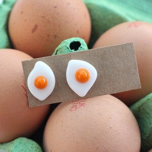 May include: A pair of white glass earrings shaped like fried eggs with orange yolks. The earrings are displayed on a brown card with the text "UK Etsy" printed on it. The card is placed on top of a carton of brown eggs.
